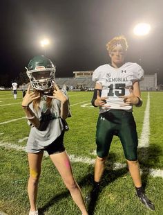 two girls in football uniforms standing on a field at night, one holding a helmet