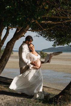 a pregnant woman in a white dress is holding her husband's belly as they stand under a tree by the water