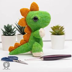 a crocheted green dragon sits next to scissors and succulents on a table