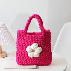 a crocheted pink purse with a flower on the front and side, sitting on a table next to a lamp