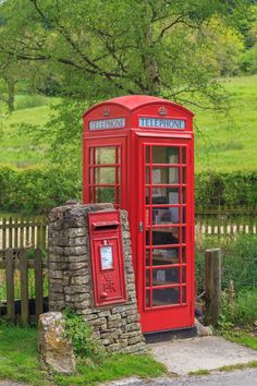 a red phone booth sitting next to a stone wall and tree in the background with green grass