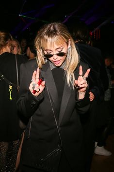 a woman in black jacket and sunglasses making the peace sign with her hands at a party