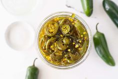 pickled peppers in a glass jar next to some green peppers on a white surface