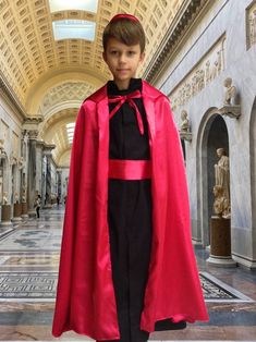 a young boy dressed in a red cape and black dress standing on a marble floor