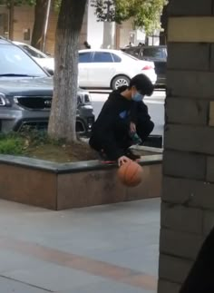 two young boys playing basketball on the sidewalk in front of parked cars and trees, with one boy holding an orange ball