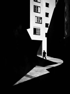 black and white photograph of person walking in an alley between two buildings with shadows on the ground