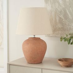 a table lamp sitting on top of a white dresser next to a bowl and painting