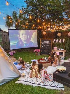 kids are sitting on the grass in front of an outdoor movie screen with lights strung over them