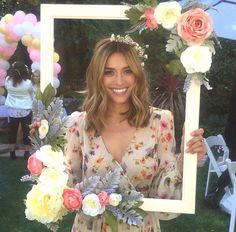 a woman standing in front of a frame with flowers and balloons around her head, holding a bouquet of flowers