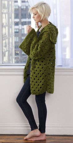 a woman standing in front of a window holding a coffee cup and wearing a green crocheted cardigan