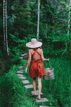 a woman wearing a straw hat walks down a path in the woods carrying a basket