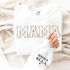 "This cozy crewneck is the perfect gift for a Mama ( or for yourself of course)!  Featuring the word \"Mama\" in bold letters on the front, stylish and cute all year wear. The shirt comes personalized with her kids names on the sleeve. The chenille letter patches are a beautiful white chenille with gold glitter backing, exuding an aura of confidence and vibrancy that's bound to catch everyone's eye. What sets them apart is the sumptuous, fluffy chenille texture that's not only pleasing to the to White T-shirt With Letter Embroidery For Gift, White T-shirt With Letter Embroidery As Gift, Cotton Sweatshirt With Lettering For Gift, White Cotton Sweater With Name Print, White Tops With Embroidered Graphics For Mother's Day, Embroidered White Tops For Mother's Day, White Embroidered Tops For Mother's Day, Mother's Day Embroidered Text Crew Neck Top, Mother's Day Crew Neck Top With Embroidered Text