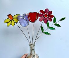 a hand holding a glass vase with colorful flowers in it on a white wall background