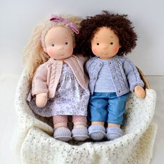 two dolls sitting next to each other in a basket on a white surface with a blanket