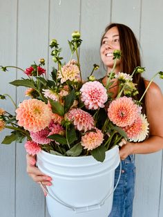 a woman holding a white bucket filled with lots of pink and orange flowers in front of a wooden wall