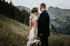 a bride and groom standing on top of a grass covered hillside with mountains in the background