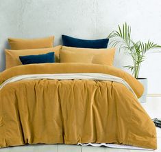 a bed with yellow sheets and blue pillows in a white room next to a potted plant