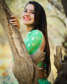 a woman in a green sari is smiling and leaning against a tree with her hand on the trunk