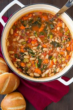 a pot filled with beans and vegetables next to rolls on a red cloth, along with a wooden spoon