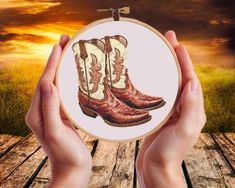 a person holding up a cross stitch picture with cowboy boots on it in front of a sunset