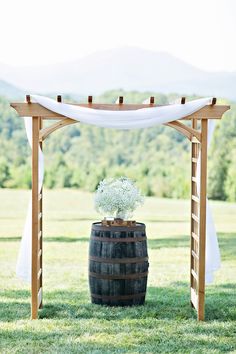 a wooden barrel with flowers in it sitting under an arbor on the grass next to a white drape