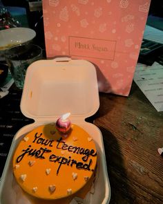 an orange cake with writing on it sitting in a white container next to a pink box