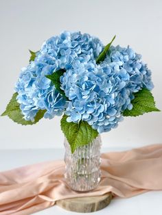 a glass vase filled with blue flowers on top of a pink table cloth next to a white wall