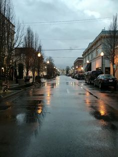 a wet street with cars parked on the side and lights hanging from the building across the street