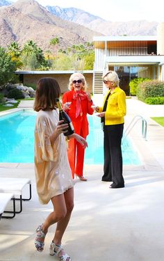 three women standing by a pool talking to each other in front of a house with mountains in the background