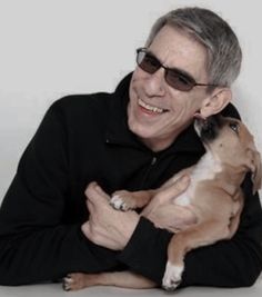 a man holding a dog in his arms and smiling at the camera while wearing sunglasses