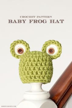 a crocheted green frog hat on top of a white vase next to a wooden stick