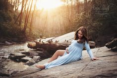 a pregnant woman sitting on top of a rock near a river in the woods at sunset