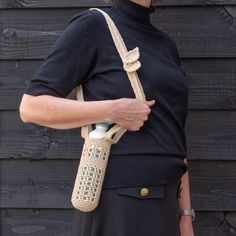 a woman is holding a crocheted bag with a bottle in her hand while standing next to a wooden wall