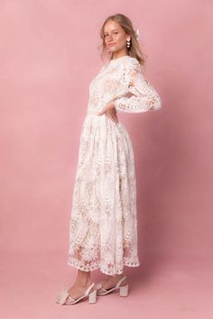 a woman wearing a white dress with long sleeves and an open back, standing against a pink background