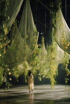 a woman standing in front of some hanging plants and flowers on a stage with sheer curtains over her head