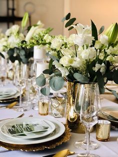 the table is set with white flowers in vases and gold place settings for dinner