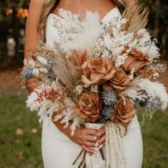 a woman holding a bouquet of dried flowers and feathers in her hands while wearing a white dress