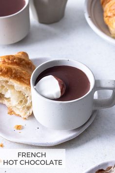 french hot chocolate in a white cup and croissant on a plate next to coffee cups