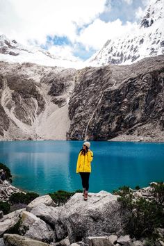 a person standing on top of a rock next to a lake with snow covered mountains in the background