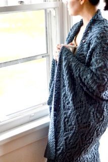 a woman standing in front of a window wearing a blue knitted cardigan sweater