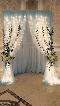 two white vases with flowers and greenery on them in front of a curtain