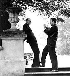 two men standing next to each other in front of a tree and stone pillar with an urn on it