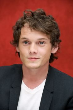 a man with curly hair wearing a black jacket and white t - shirt is looking at the camera