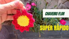 a hand holding a crocheted flower in front of some pink and yellow flowers