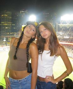 two women standing next to each other in front of a baseball field with lights on