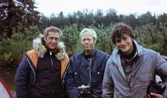 three men standing next to each other in front of a river with trees behind them