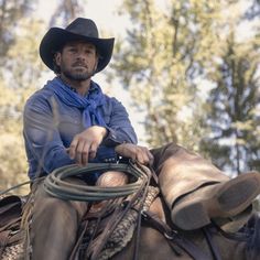 a man wearing a cowboy hat riding on the back of a brown horse in front of trees