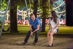 a man and woman sitting on swings in a park at night with lights behind them