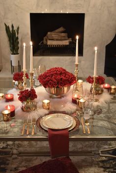 a glass table topped with red roses and gold vases filled with candles next to a fire place