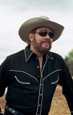 a man wearing a cowboy hat and sunglasses standing in front of a body of water